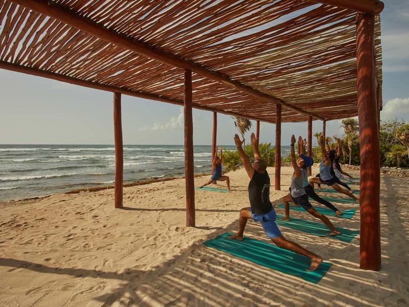 Persone che praticano yoga su tappetini sotto un padiglione di legno su una spiaggia sabbiosa con vista sul mare.