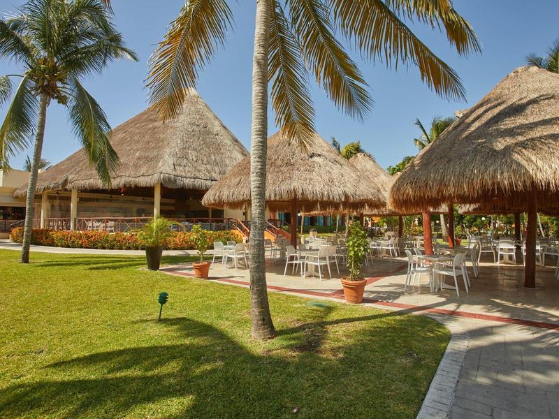 Palme e padiglioni con tetto di paglia accanto a un prato ben curato in un ambiente vacanziero tropicale.