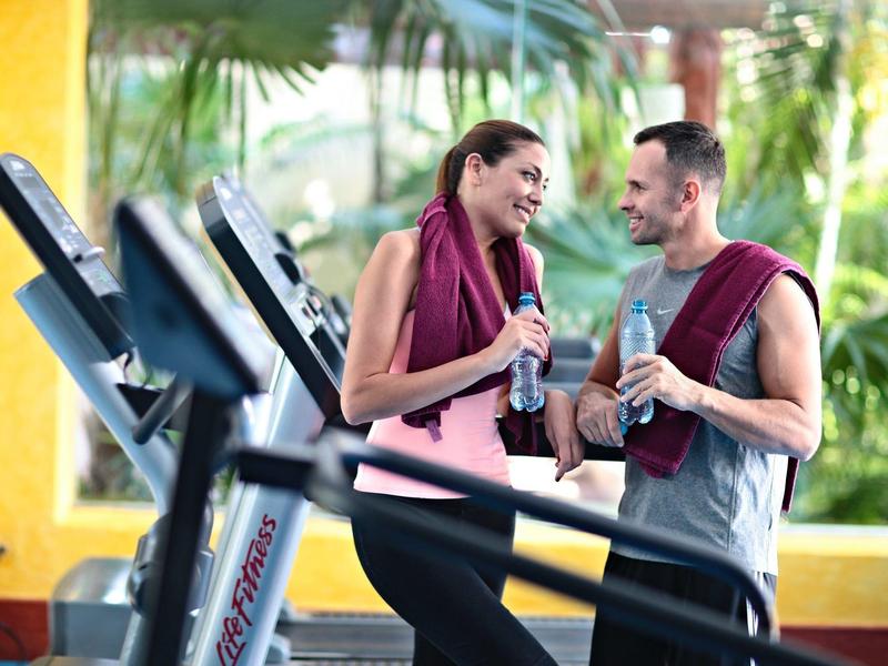 Coppia che parla e beve acqua in palestra con tapis roulant e decorazioni tropicali.