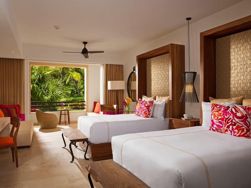 Camera d'hotel moderna con due letti, cuscini colorati e vista su piante tropicali.