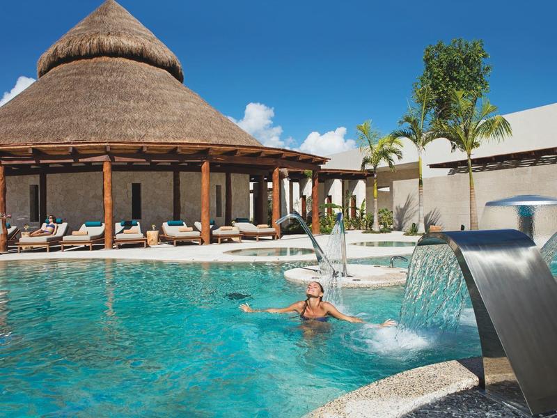 Piscina con cascata e palme davanti a un padiglione rotondo in un resort sotto un cielo azzurro.