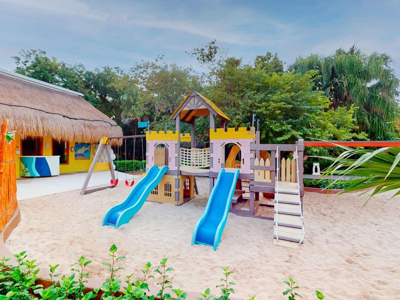 Area giochi per bambini con scivoli e altalena su sabbia in un giardino.