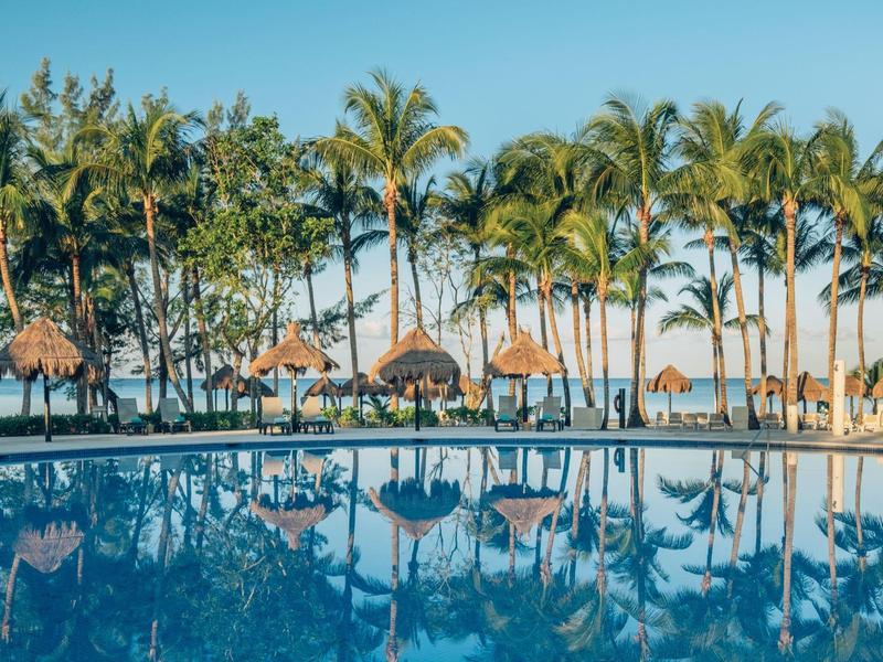 Una piscina con acqua riflettente davanti a palme e ombrelloni di paglia sulla spiaggia.
