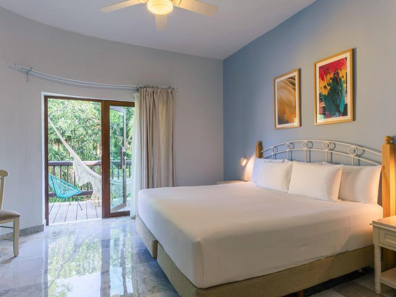 Camera d'hotel luminosa con letto grande, porta scorrevole per balcone e vista su piante verdi.