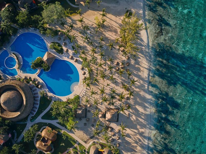 Veduta aerea di un hotel con piscina e palme accanto a una spiaggia turchese.