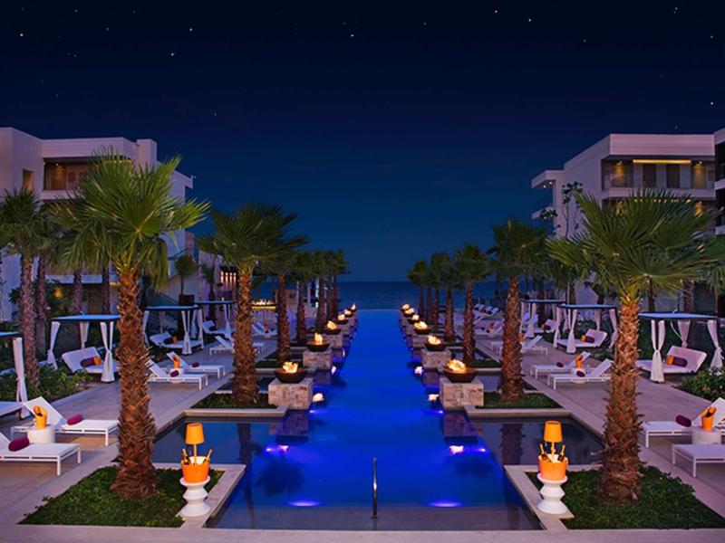 Vista notturna di una piscina esterna illuminata con palme e lettini in un resort.