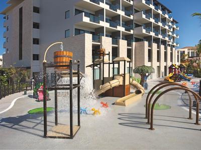 Area giochi moderna con scivoli e strutture per arrampicata vicino a un edificio alberghiero multipiano sotto un cielo sereno.