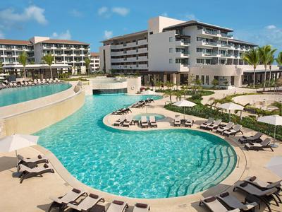 Hotel moderno con grande piscina curva e lettini al sole sotto un cielo blu.