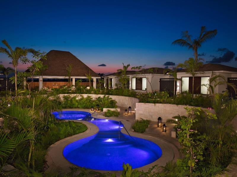 Vista notturna di una piscina curva illuminata circondata da piante tropicali e bungalow.