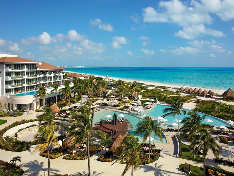 Resort con piscine, palme e spiaggia sotto un cielo azzurro con nuvole.