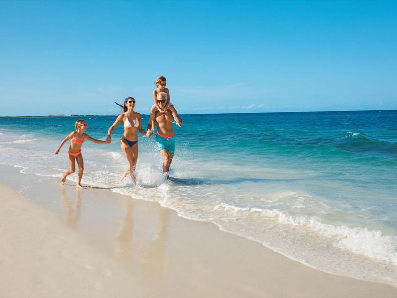 Famiglia che corre e gioca su una spiaggia soleggiata con cielo azzurro e onde del mare.