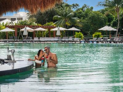 Due persone si rilassano in una grande piscina con ombrelloni e palme sullo sfondo.