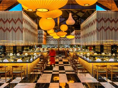 Moderní restaurace se žlutými lampami, dřevěným stropem a černobílou šachovnicovou podlahou.