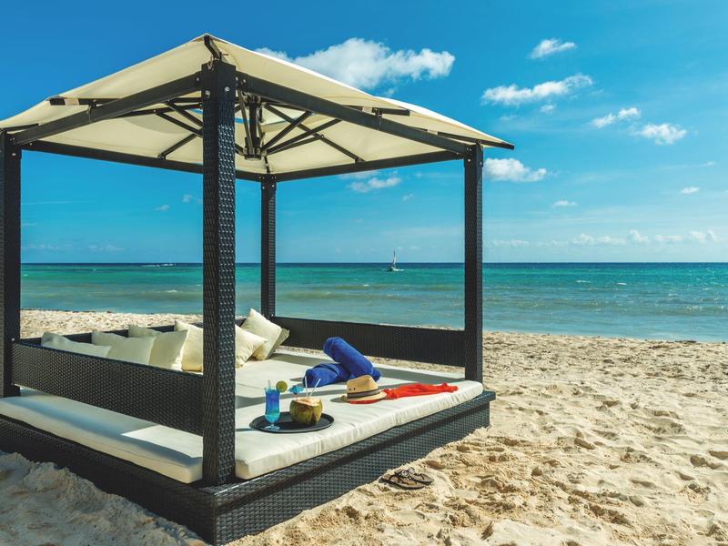 Un lussuoso lettino da spiaggia ombreggiato con cuscini e bevande sulla sabbia vicino al mare.