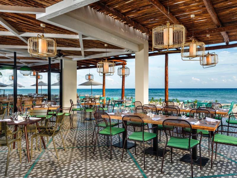 Ristorante all'aperto con vista mare, soffitto in legno e tavoli rotondi con sedie verdi.