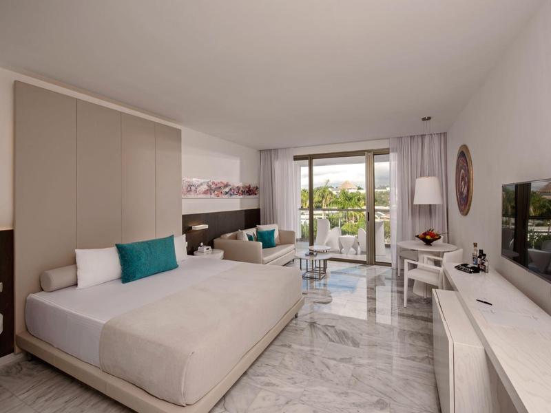 Chambre d'hôtel lumineuse avec lit double, coin salon et balcon donnant sur la verdure.