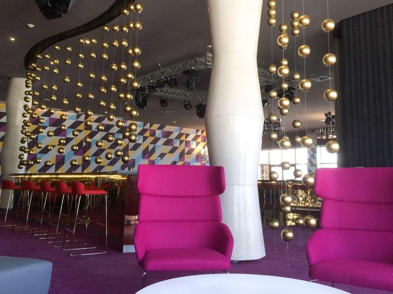 Hall d'hôtel moderne avec fauteuils violets, luminaires suspendus sphériques et tabourets colorés.