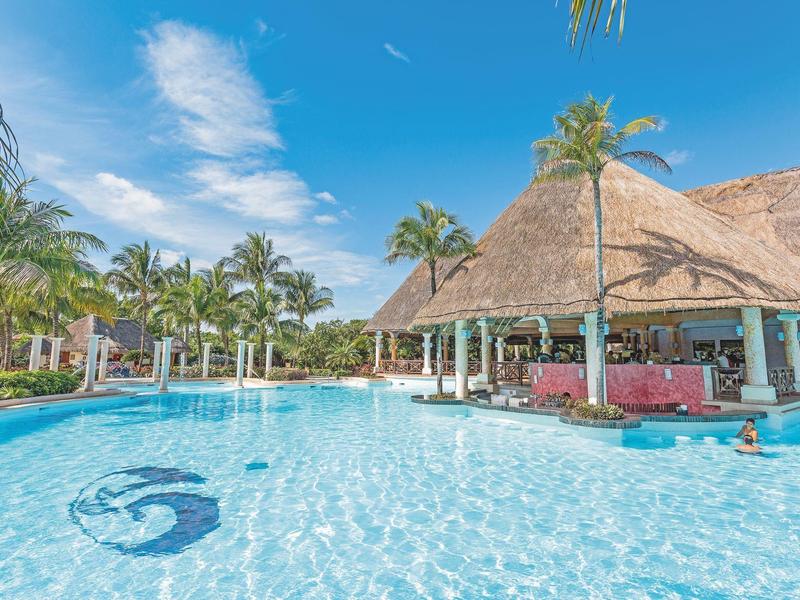Piscina azzurra con bar in acqua e palme sotto un cielo azzurro.