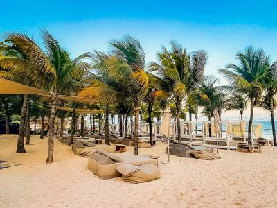 Strand mit Palmen, Sand und Liegestühlen unter Sonnenschirmen bei blauem Himmel.