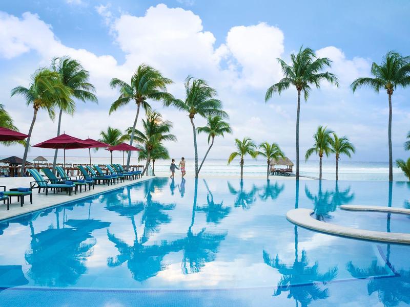 Una piscina che riflette palme e ombrelloni sotto un cielo sereno in spiaggia.