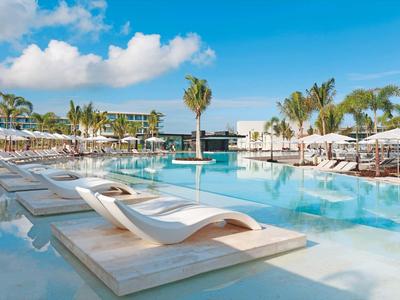 Moderní bazén s lehátky a palmami v hotelovém resortu pod modrou oblohou.