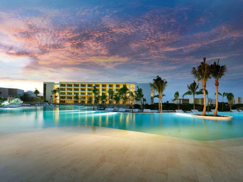 Un hotel moderno con piscina illuminata e palme sotto un cielo serale colorato.