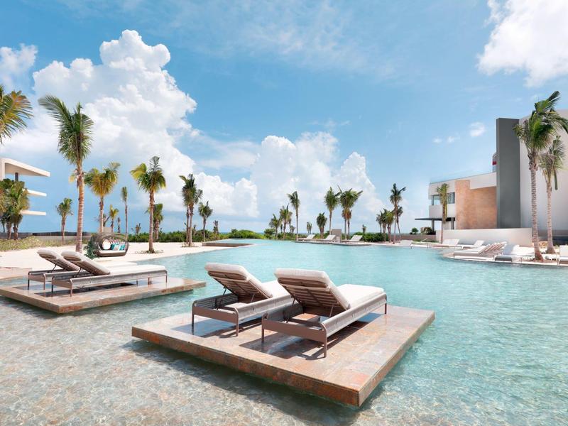 Piscina moderna dell'hotel con lettini su piattaforme di legno, palme e cielo azzurro.