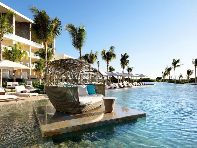 Hotel di lusso con piscina, lettini e un moderno spazio salotto sotto un cielo azzurro brillante.
