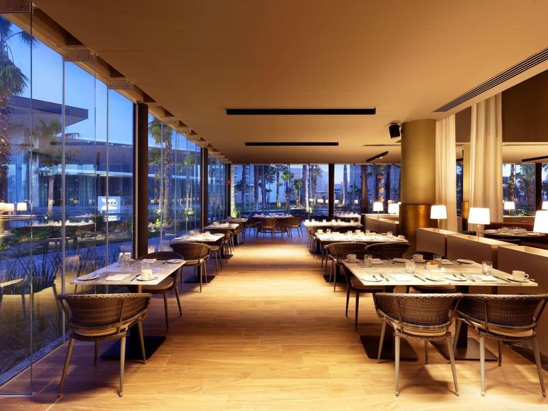 Area ristorante moderna dell'hotel con pareti di vetro, pavimenti in legno e tavoli elegantemente apparecchiati.