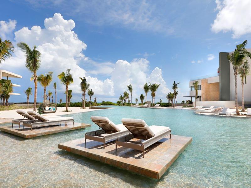 Complesso alberghiero moderno con piscina e lettini prendisole sotto un cielo blu.