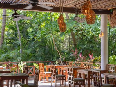 Überdachter Außenbereich eines Restaurants mit Holztischen und -stühlen, umgeben von üppigem Grün.