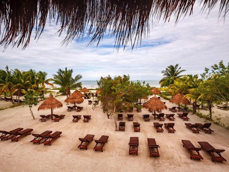 Plage avec de nombreuses chaises longues et toits en chaume sous un ciel bleu et des palmiers.