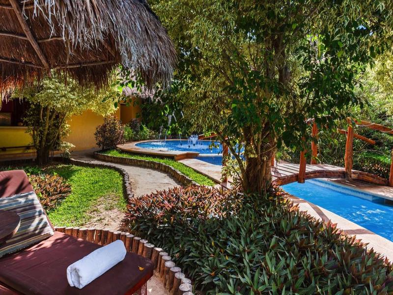 Espace piscine avec chaises longues, pelouses et un pont en bois dans un jardin tropical.
