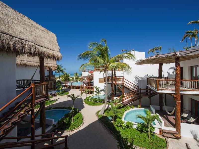 Un complexe hôtelier ensoleillé avec des bâtiments blancs, des terrasses en bois et une végétation tropicale sous un ciel bleu.