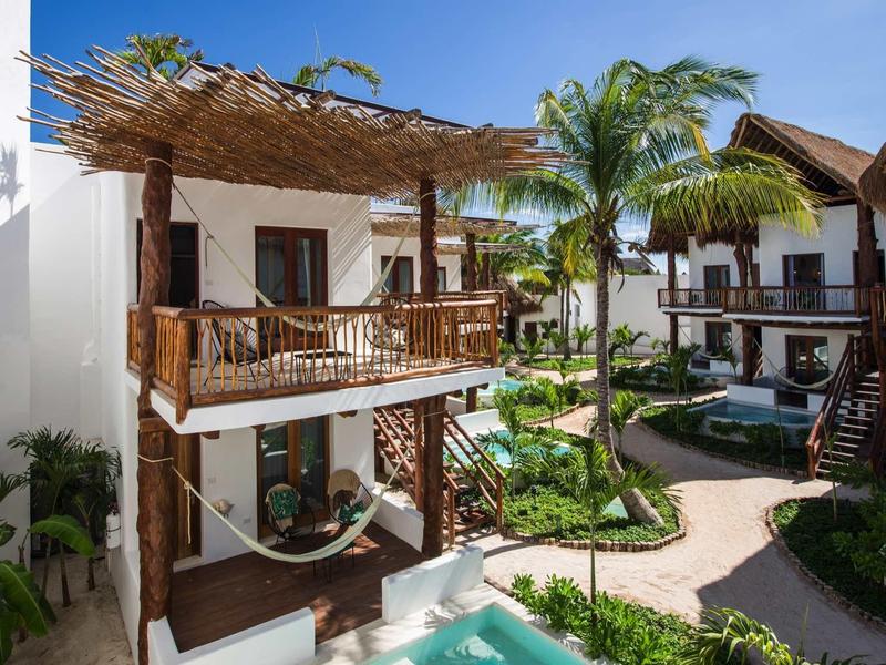 Hôtel moderne à deux étages avec balcons, palmiers et petite piscine dans un cadre tropical.