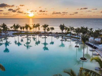 Großer Pool mit Palmen am Meer, Sonnenuntergang und leicht bewölktem Himmel.