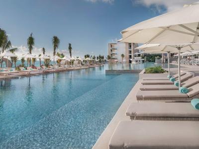 Moderner Poolbereich mit Liegen und weißen Sonnenschirmen, Palmen und blauem Himmel im Hintergrund.