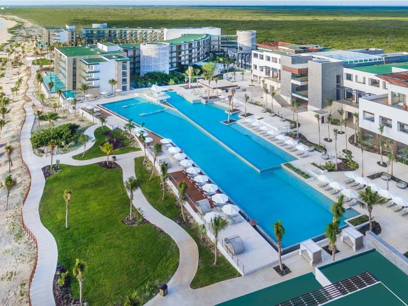 Moderne Hotelanlage mit großem blauen Swimmingpool, Palmen, weißen Gebäuden und Strand am Meer.