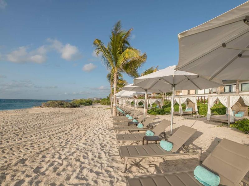 Sandstrand mit Palmen, Liegen und Sonnenschirmen vor klarem blauem Himmel und ruhigem Meer.