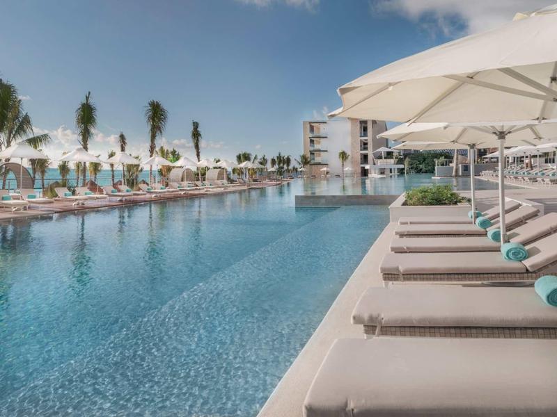 Moderner Poolbereich mit Liegen und weißen Sonnenschirmen, Palmen und blauem Himmel im Hintergrund.