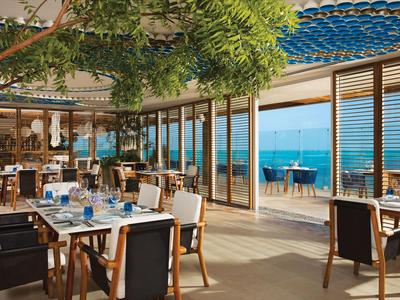 Helles Restaurant mit modernem Design, Holzstühlen und Blick auf das Meer durch große Fenster.