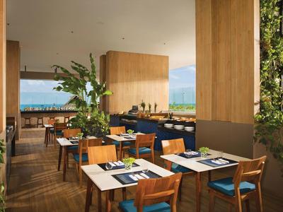 Modernes Restaurant mit Holzmöbeln, grünen Pflanzen und Blick aufs Meer durch große Fenster.