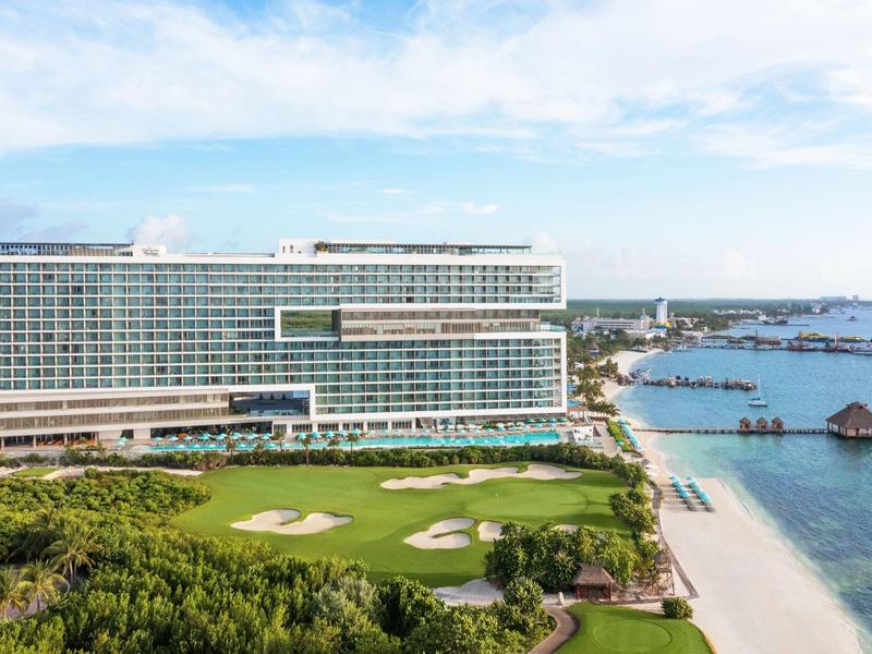 Bâtiment d'hôtel moderne avec terrain de golf et plage au bord de la mer sous un ciel bleu.