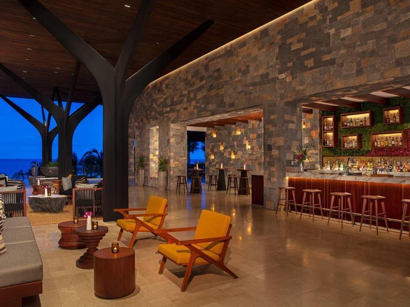 Area bar aperta dell'hotel con comode poltrone, parete in pietra e illuminazione serale vicino al mare