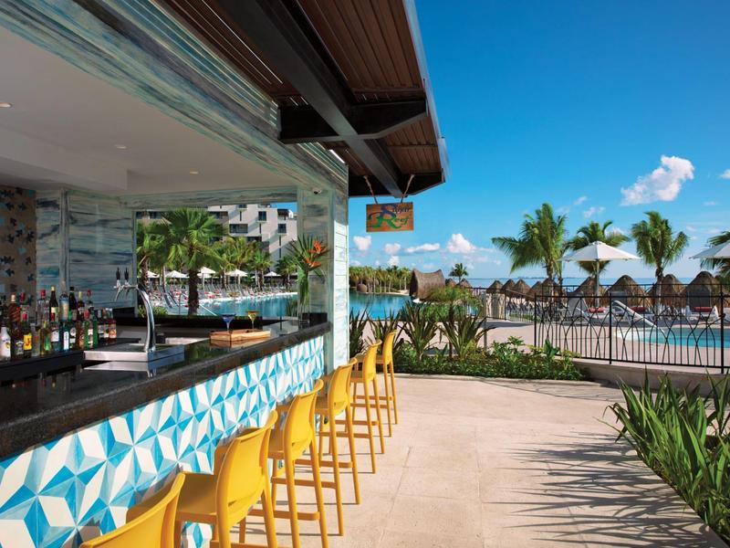 Bar all'aperto con sgabelli gialli che si affaccia su una piscina con palme sotto un cielo sereno.