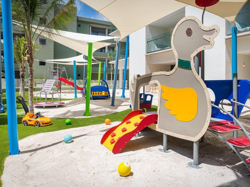 Zona de juegos infantiles con tobogán y columpio junto a un hotel con toldos en el exterior.