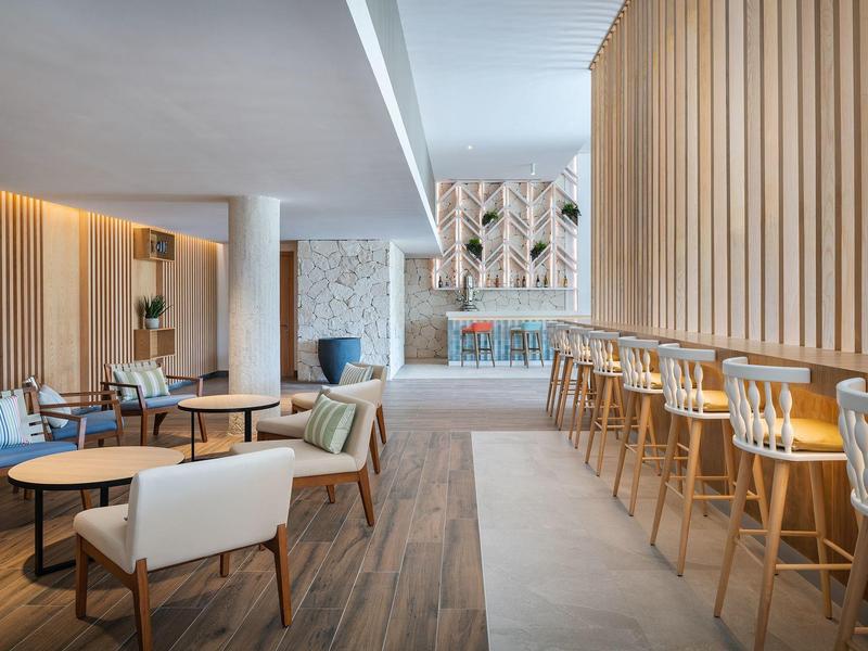 Recepción moderna con barra de madera, sillas de diseño y área de descanso luminosa en hotel.