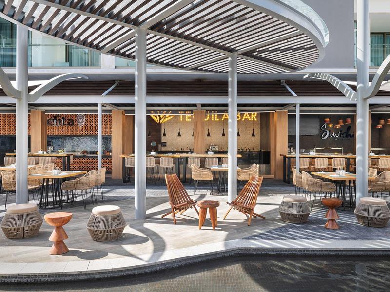 Área exterior moderna con asientos y mesas bajo una pérgola curva junto al agua.