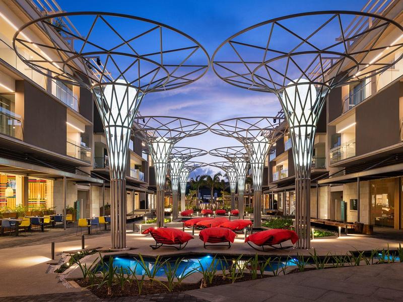 Área exterior moderna de un hotel con luces decorativas iluminadas y muebles rojos al anochecer.