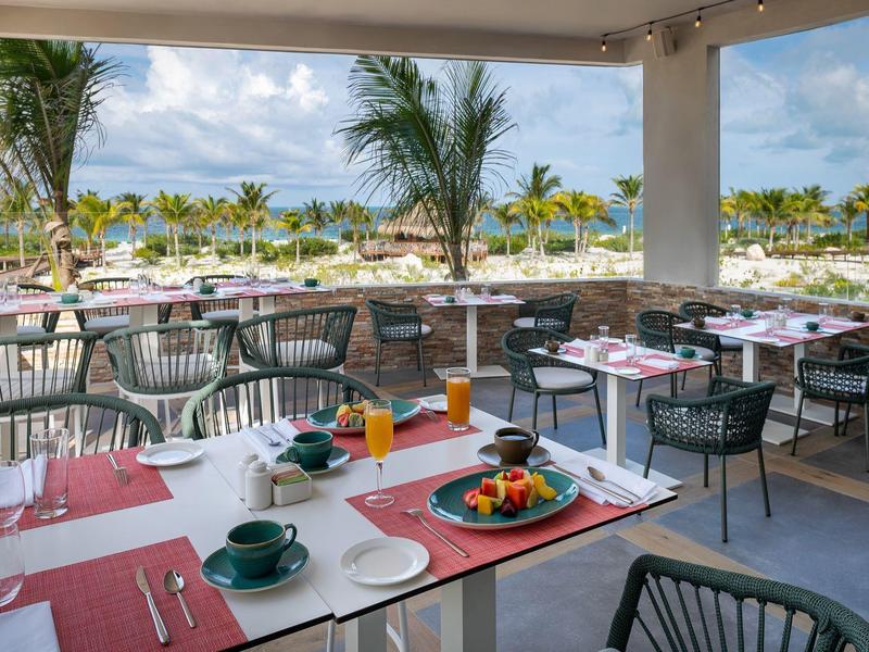 Terraza de restaurante con mesas y sillas, vista a palmeras y al mar en un hotel tropical.